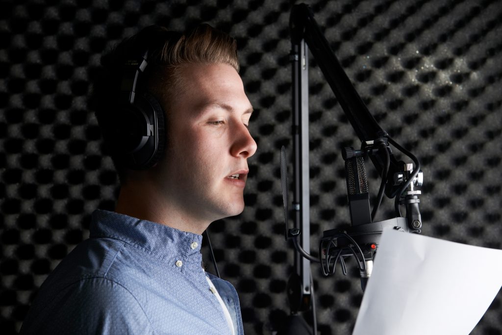 man in sound booth recording studio wearing headphones and reading from script into microphone for professionally recorded audio and voicing