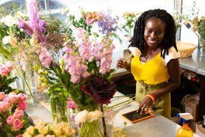 Cheerful dark-skinned girl standing at her workplace and enjoying fresh flowers - overhead marketing concept where employee is enjoying their work