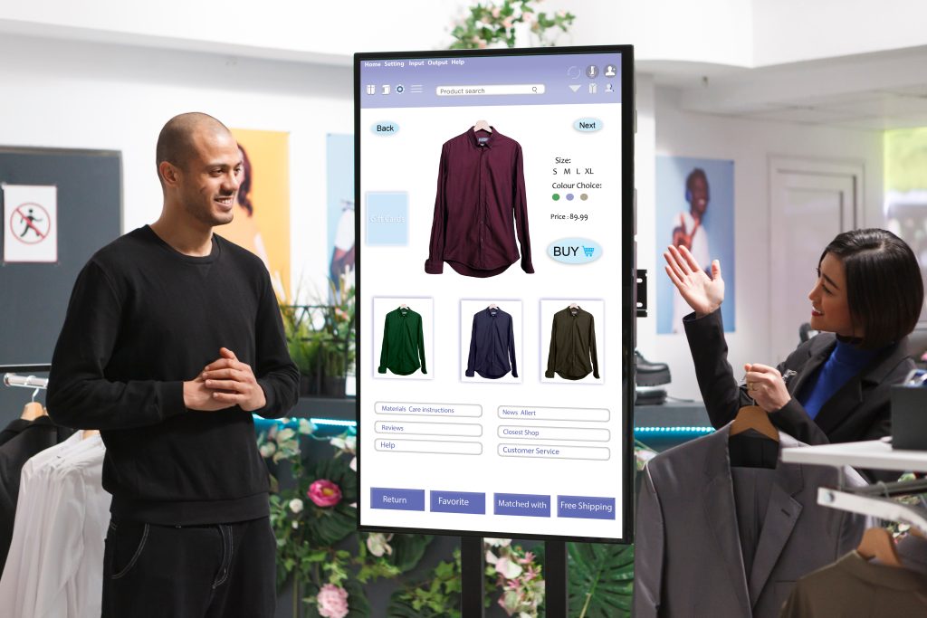 Digital signage is more than just digital posters; it's a versatile and powerful communication tool that, when implemented effectively, can enhance the customer experience, increase sales, and provide valuable insights into customer behavior - male customer and female store associate discussing clothing shown on a digital screen in well lit clothing store 