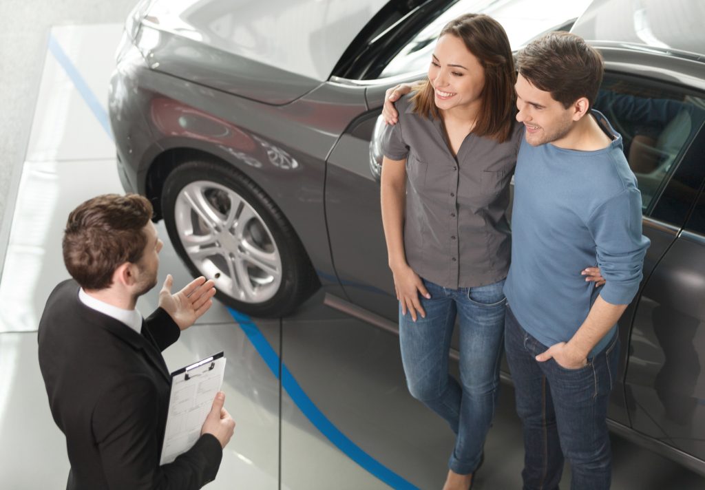 Strategic Overhead Music and Messaging in Car Dealerships - background music and overhead marketing concept - smiling couple speaking with car salesman at dealership standing in front of a new car