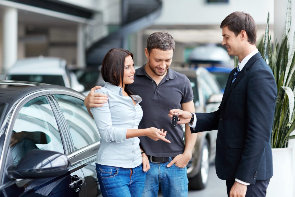 The strategic use of overhead music and messaging in car dealerships can transform the customer experience, making it more engaging and enjoyable - overhead marketing concept - couple smiling and taking keys from salesman in care dealership showroom showing a completed sale of a new car