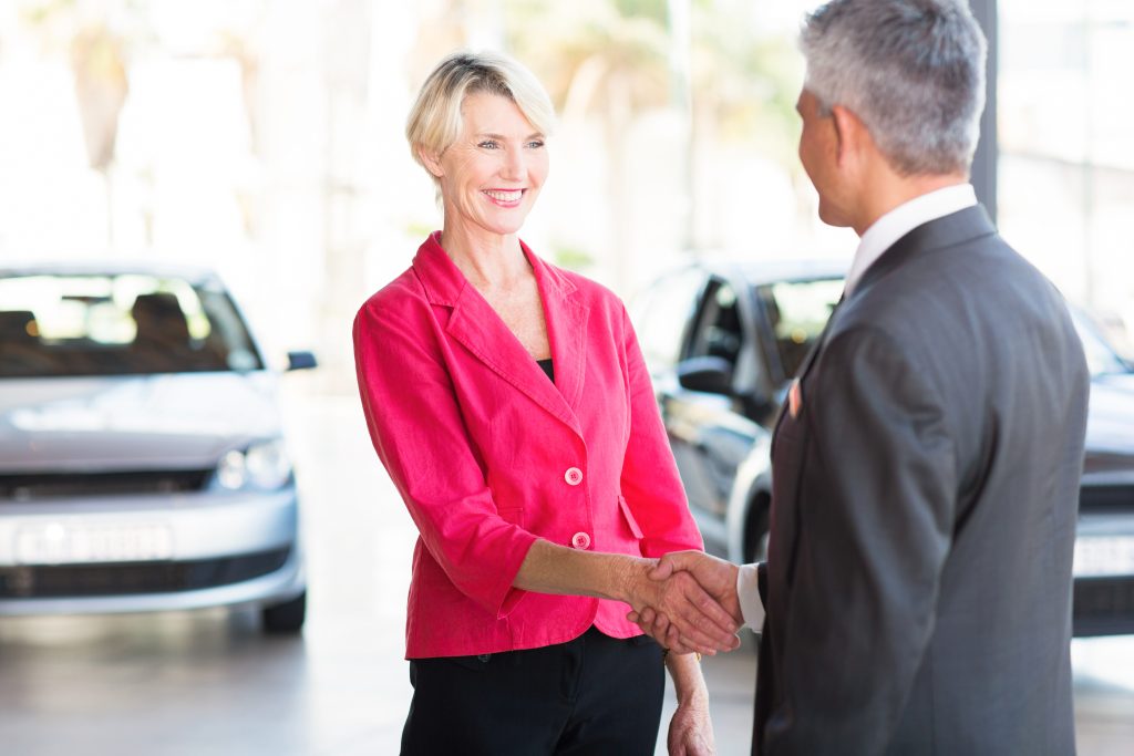 Leveraging Overhead Messaging to Enhance Sales and Service - background music and overhead marketing concept - salesman shaking hands with smiling woman in car dealership show room showing positive sales interaction of buying a new car