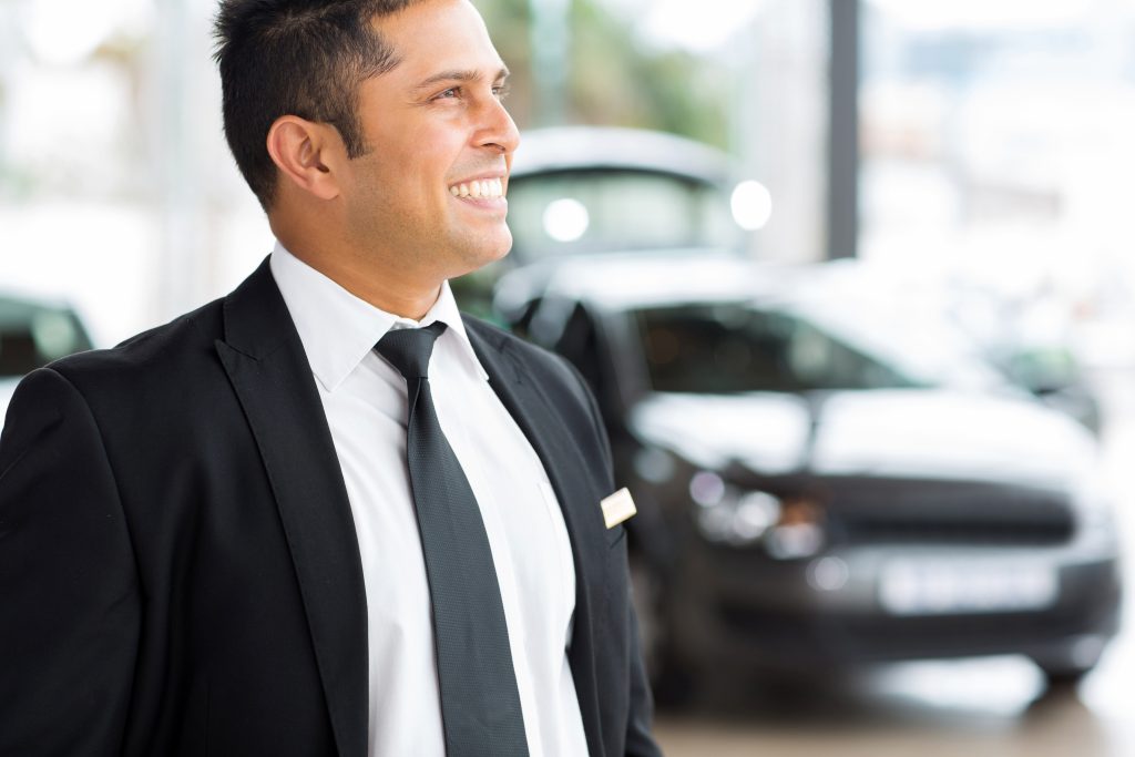 The Role of Overhead Music in Car Dealerships - Overhead marketing and background music and messaging in car dealership concept - smiling salesman standing in front of new cars in dealership showroom