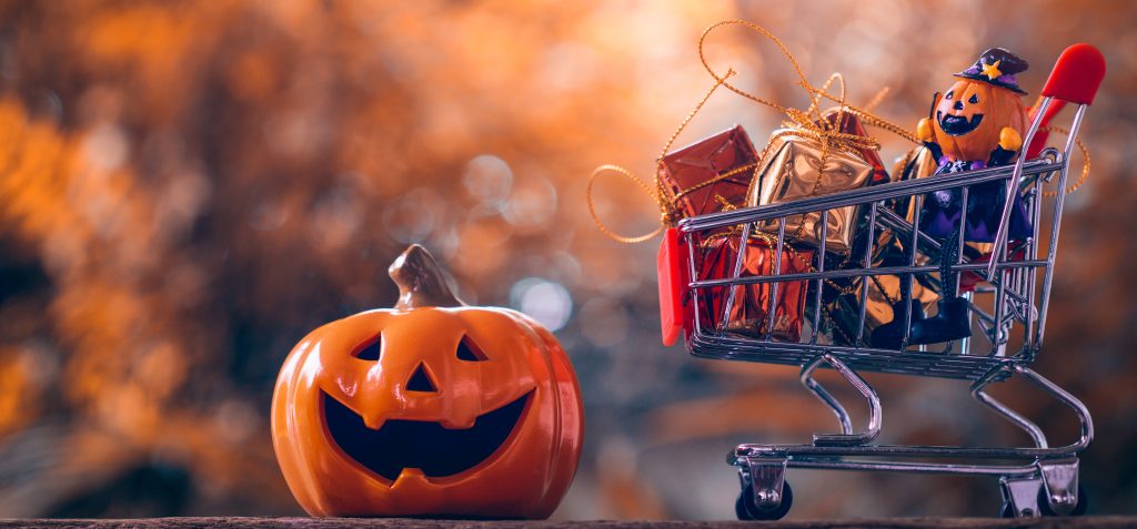 Halloween shopping image - miniature shopping cart filled with mini gifts and a jack-o-lantern character next to a larger smiling jack-o-lantern pumpkin against a blurred orange and black background