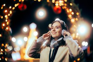 woman standing outside amongst Christmas lights and decorations with headphones on and a calm happy expression on her face, listening to holiday music at a holiday event